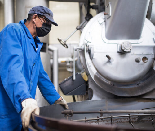 Worker in face mask controlling coffee roasting process. Coffee roaster working on roasting equipment. Man in mask and uniform Working with machinery appliance. Production, food concept