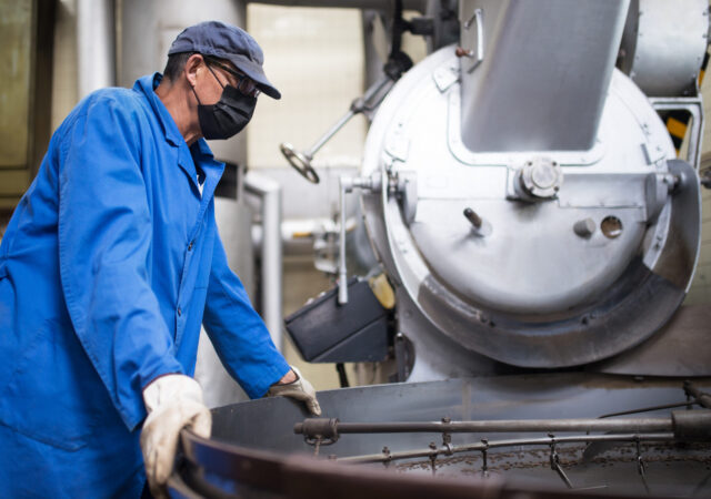 Worker in face mask controlling coffee roasting process. Coffee roaster working on roasting equipment. Man in mask and uniform Working with machinery appliance. Production, food concept