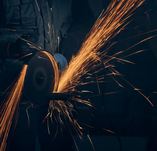 Close up of worker in special black suit working with angle grinder and grinding metal with large yellow sparks. Concept of polishing metal with special equipment in dark room.
