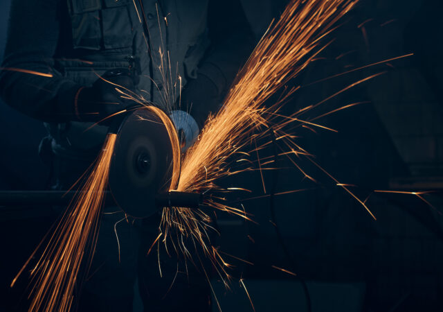 Close up of worker in special black suit working with angle grinder and grinding metal with large yellow sparks. Concept of polishing metal with special equipment in dark room.